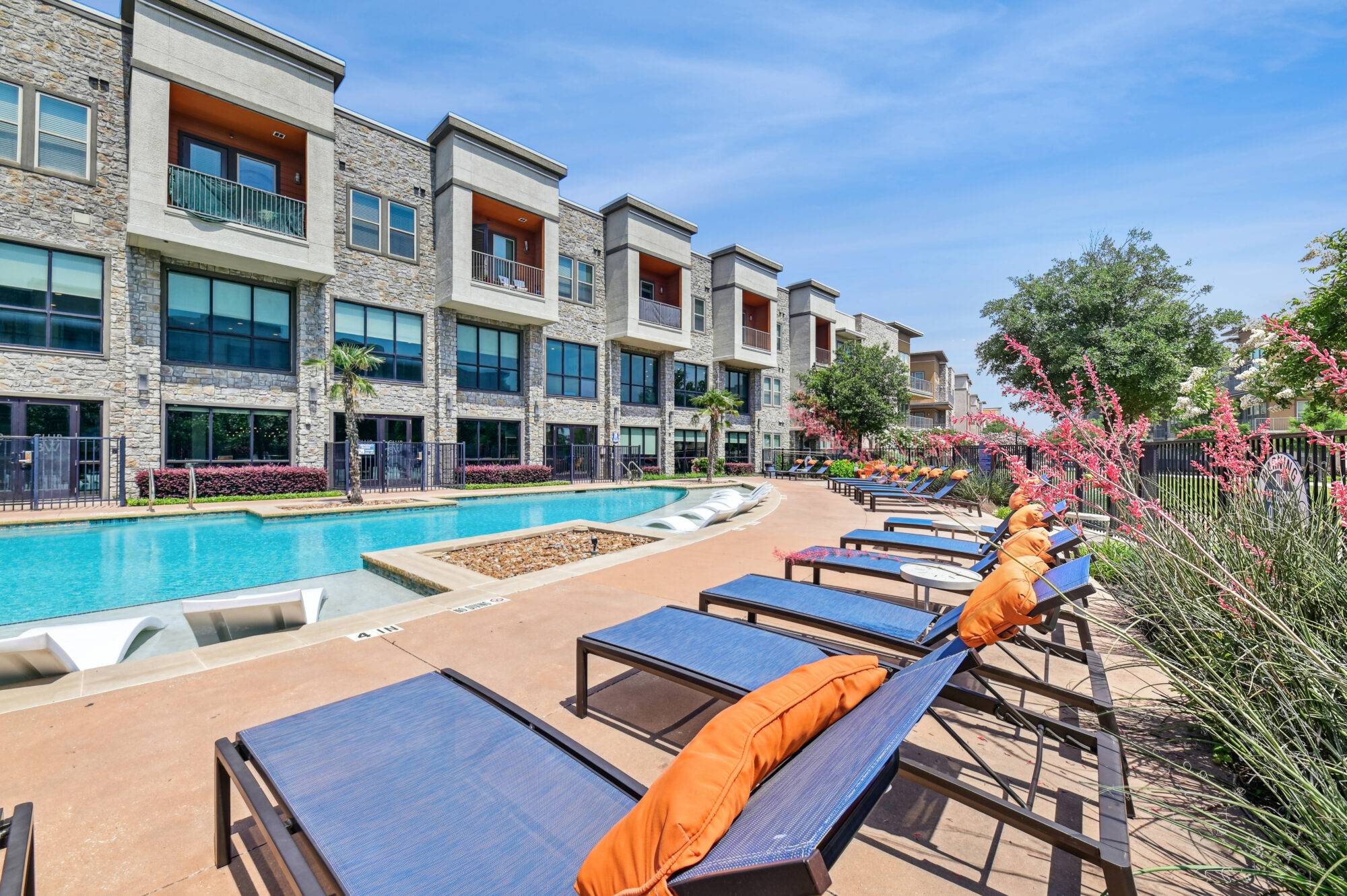 Modern apartment complex with a swimming pool, lounge chairs with orange cushions, and flowering plants along the poolside under a sunny blue sky. Balconies overlook the pool area.